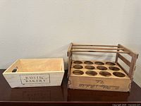 Two wooden items side by side: a light Rustic Bakery crate and a darker vintage Rothschild Cellar wine bottle carrier with bottle slots.