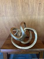 Front view of metal infinity knot sculpture showing intertwined loops and textured gold/brass finish on wooden base.