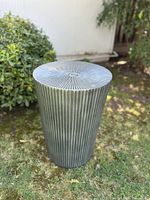 Full view of vintage iron side table with ribbed vertical texture on the base, photographed outside on grass with green shrubbery in background.