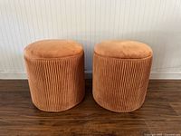 Front view of two orange velvet ribbed pouf ottomans side by side on wood floor against light wall.