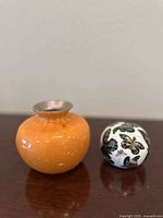 Image showing front view of the small glossy orange porcelain vase and the butterfly-decorated porcelain ball side by side on a wooden surface.