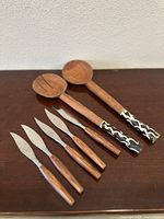 Full view of the four knives and two serving utensils arranged horizontally on a wooden surface against a white wall.