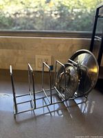 Photo of a vintage stainless steel drying rack placed on a kitchen counter with a window in the background, showing four vertical slots and two pot lids stored in the rack.