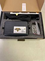 Opened box showing rectangular black Auxbeam control unit with ridge design, braided wiring cable, and bag of screws for mounting.