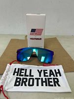 Front view of sunglasses with blue mirrored lens and white box with Cleetus x Heatwave branding, placed behind. Microfiber pouch with 'HELL YEAH BROTHER' text in front.
