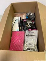 Open cardboard box showing assorted electronic items including screen protector, chargers, cables, and a pink quilted case