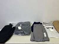 Overview photo of 8 clothing items neatly folded or in packaging, showing variety of colors: black, grey, beige, striped, and white with text.