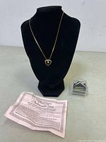 Gold plated necklace with heart-shaped pendant displayed on black velvet necklace stand. Certificate and small box beside the stand.