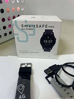 Photo showing SmartSAFE Pro packaging box with watch face image, watch with black strap, and USB charging cable.