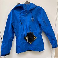 Front view showing Macaw blue shell, central waterproof zipper, Arc’teryx logo and attached tag