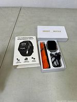 Photo showing the open box containing a smartwatch, an orange strap, a black strap, USB charging cable, and the user manual which highlights calling, health, fitness apps, and additional functions.