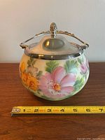 Front view of milk glass biscuit barrel with floral hand painting and silver plated lid and handle, with measuring tape showing approximately 7 inches diameter.