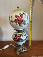 Front view of antique electrified parlor oil lamp showing floral hand-painted globes and ornate brass base with measuring tape for size reference.