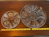 Photo shows two round sectioned serving dishes on a wooden surface with a measuring tape indicating their sizes. Left dish is smaller with pinwheel design and scalloped edge, right dish is larger with foliage pattern.