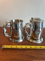Side-by-side image of two pewter beer steins on a wooden surface with ruler measuring their width. The stein on the left has a naked woman handle and engraving. The stein on the right is lidded with multiple dents.