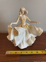 Front view of the Royal Doulton Sweet Seventeen porcelain figurine showing the detailed gown, arms extended gracefully, and facial features. A measuring tape indicates height around 7.25 inches.
