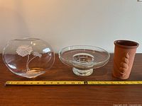 Photo showing the three items side by side on a wooden surface with a tape measure for scale: crystal vase with etched flowers, glass centrepiece bowl, and terracotta forcing vase.