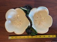 Top view of yellow ceramic morning glory shaped candy nut dish with green leaf and stem handle. Tape measure shows about 10 inches length. Small chip visible on one petal.