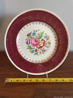 Photo of front of plate showing floral design with gold accents and maroon border with detailed gold pattern, measuring approximately 11 inches.