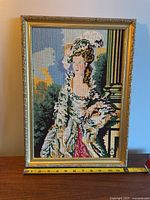 Full view of the framed vintage cross stitch portrait of a woman, featuring a gold-tone decorative frame with size dimensions shown by a ruler.