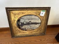 Frontal view of the oval black and white coastal painting in an ornate gilded frame, showing full artwork and frame detail.