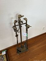 Full view of brass fireplace tool set with duck head handles and stand on wooden floor.