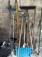 Full view of gardening tools leaning against wall including rakes, shovels, snow shovel and broom.