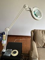 Magnifying bench lamp mounted on a table, adjustable white arm extended, lamp head with circular magnifying glass, additional lens and magnifier shown on table.