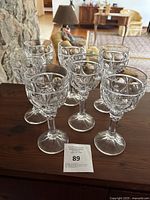 Eight clear crystal wine goblets arranged on wooden surface with natural room background.