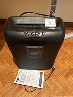Front view showing Staples paper shredder with control panel, paper slot, power cord, and instruction manual on floor
