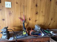 Power tools including an orbital palm sander, Ryobi hammer drill, staple gun, and various hand tools on wooden shelf against paneling wall.