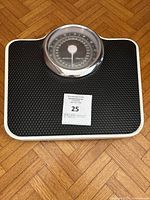 Top down photo of a black textured platform mechanical bathroom scale with a large circular analog dial and a chrome bezel resting on wooden parquet flooring.