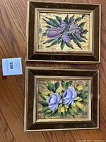 Two framed floral paintings, each with purple flowers and green leaves on a gold-textured background, displayed side by side on a wooden floor