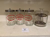 Front view of three labeled vintage glass jars (Cotton, Tongue Depressors, Bandages) with metal lids placed next to a clear glass instrument jar with visible metal insert.
