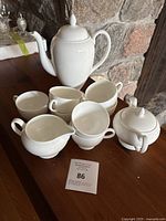 Photo showing the assembled tea set with teapot, creamer, sugar bowl, 5 cups stacked, and plates underneath on wooden surface by a stone wall.