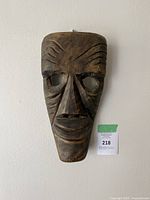 Front photo of a carved wooden African mask mounted on a wall, showing eye holes and detailed facial carvings.