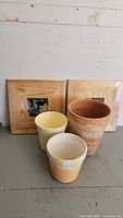 Photo showing three flower pots and two wooden picture frames arranged on a flat surface against a white wall.