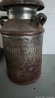 Photo shows side of metal milk can with embossed text 'DAIRY JARVIS' and rust on lower half.