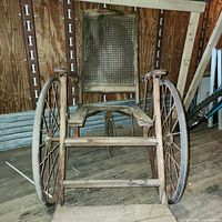 Front view of the antique wheelchair showing large wooden wheels, wooden frame, and cane backrest. The seat section is missing the cane surface.