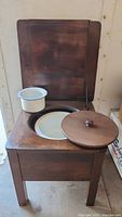 Full view of antique wooden chamber pot with hinged lid open, showing interior and one enamel pot placed on top.
