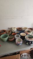 Photo showing a large assortment of over 20 planter pots and pots with clay bottoms of various sizes, styles and colors arranged on a table.