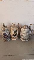 Three ceramic collectible beer steins with pewter lids displayed side-by-side on a flat surface.