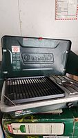 Open Coleman propane camping stove showing two burners and metal grates with green lid displaying Coleman logo and safety labels.