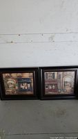 Pair of framed European street scene art prints on a floor against white wood paneling, showing both pieces side by side.