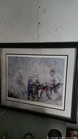 Photo showing a black wooden framed art print of a hockey scene with players on ice, signed by the artist, mounted on a wall.