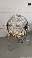 Metal bingo cage on a stand holding multiple small white numbered balls inside.