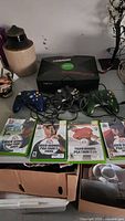 Front view of the original Xbox console with logo, three wired controllers (black, green translucent, blue), and four Tiger Woods PGA Tour games in cases.