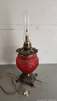 Full view of the ornate vintage electric lamp showing red glass base with floral design, brass metal base, clear glass chimney, and power cord with plug.