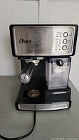 Front view of Oster PrimaLatte espresso and cappuccino machine showing control panel, portafilter, steam wand, and milk container.