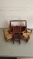 Two light wood miniature rocking chairs with woven seats, one dark reddish-brown miniature rocking chair, and a rectangular woven basket with handle placed on gray surface.